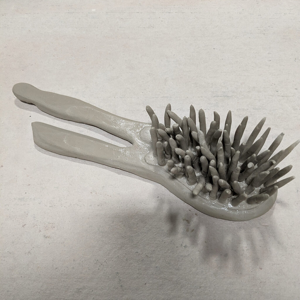 A surreal looking hairbrush with a forked handle made from gray clay.