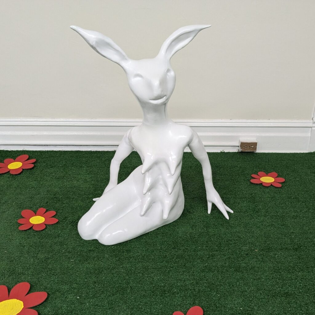 A white bunny-woman sculpture sitting in a green field with cartoon red and yellow flowers.